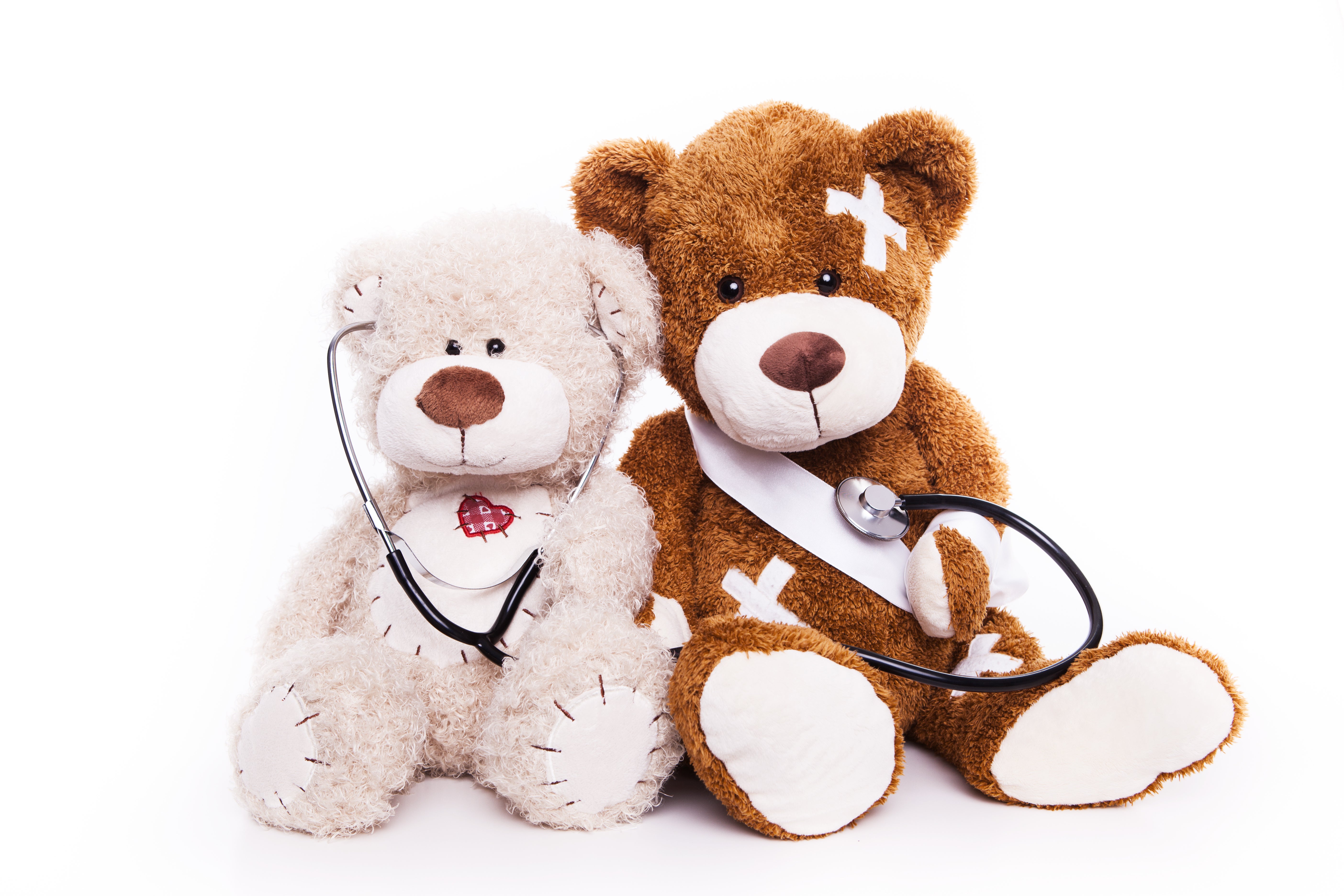 Zwei Teddybaeren sitzen nebeneinander. Einer von beiden hat Pflaster (Verletzungen) und der andere hoert ihn mit dem Stethoskop ab.